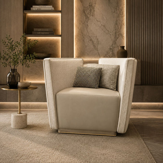 A modern beige armchair with a textured fabric upholstery, two accent pillows, and a slim gold metal base, set in a luxurious, warmly lit living room with a marble accent wall and built-in shelves.