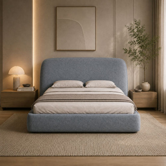 A modern platform bed with a tall upholstered headboard in a muted blue-gray fabric, styled in a warm, minimalist bedroom with a beige rug, wooden nightstands, a lit table lamp, and a potted plant.
